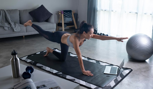 Mujer asiática joven deportiva entablado frente al espacio de copia de la vista lateral interior de la sala de estar de la computadora portátil Joven saludable en ropa deportiva haciendo ejercicio en casa viendo la clase de fitness en línea