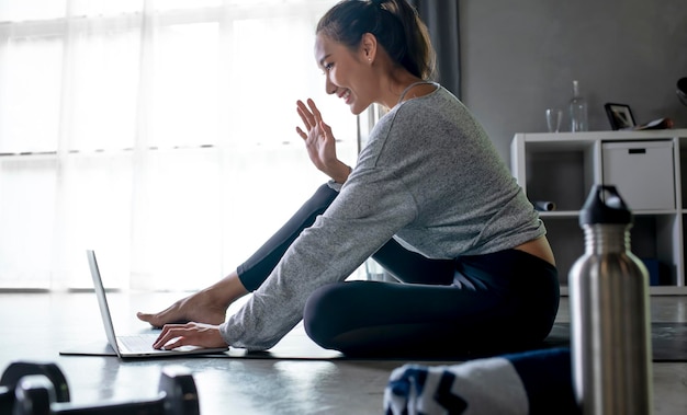 Mujer asiática joven deportiva haciendo ejercicio en casa viendo videos de fitness en Internet o teniendo clases de fitness en línea usando el espacio interior de la sala de estar de la computadora portátil