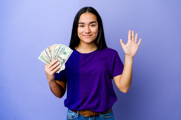 Mujer asiática joven con dinero aislado en la pared púrpura sonriendo alegre mostrando el número cinco con los dedos.