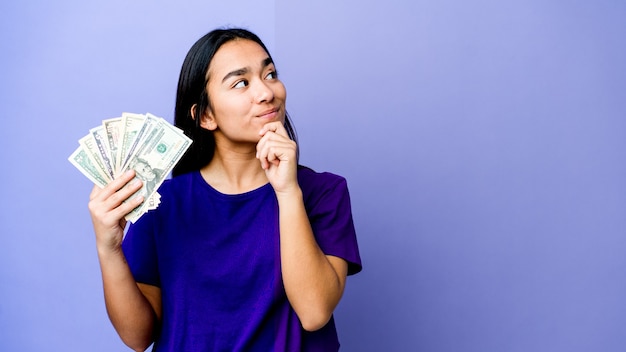 Mujer asiática joven con dinero aislado en púrpura mirando hacia los lados con expresión dudosa y escéptica.