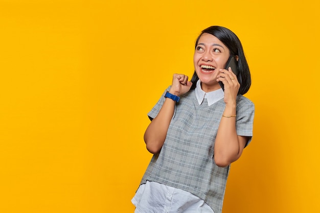 Mujer asiática joven emocionada que recibe la llamada entrante en el teléfono inteligente y que mira a un lado sobre fondo amarillo