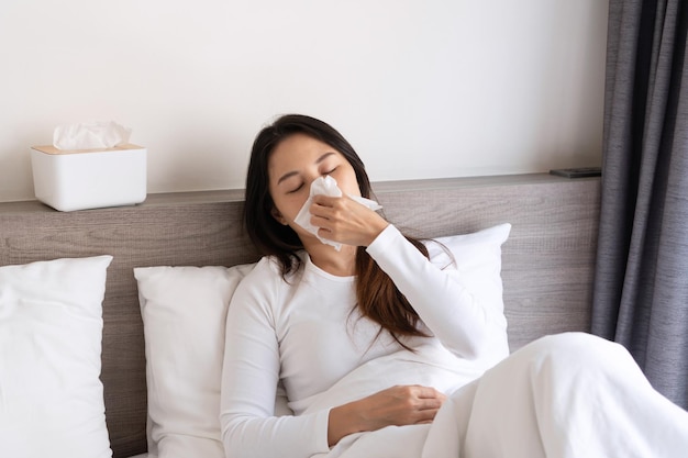 Mujer asiática joven enferma que siente frío y tiene fiebre alta sonándose la nariz mientras está acostada en la cama por la mañana en casa Concepto de gripe alérgica