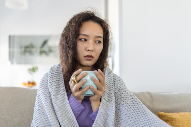 Mujer asiática joven enferma sentada en casa en la cama con una taza de té caliente y un pañuelo Resfriados estacionales tos secreción nasal infecciones virales tratamiento en el hogar
