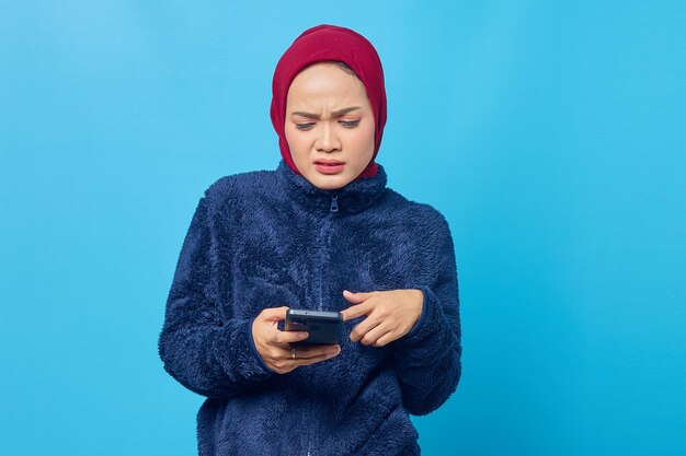 Mujer asiática joven enojada que usa el teléfono móvil y que mira la pantalla del teléfono inteligente sobre fondo azul