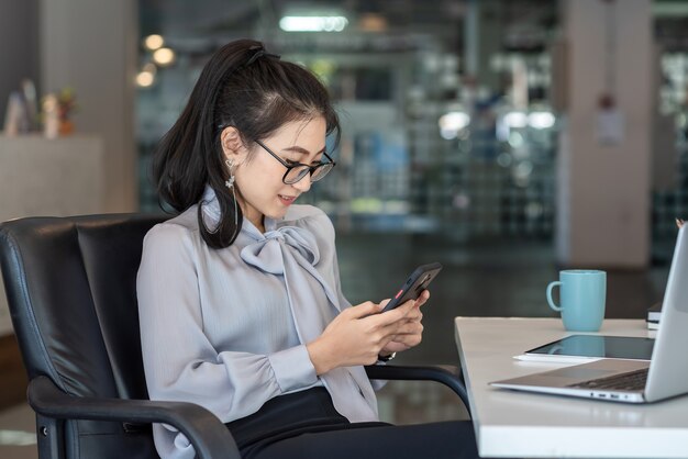 La mujer asiática joven está feliz de sentarse en el teléfono en el escritorio de la oficina.