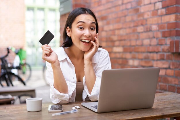 Mujer asiática joven con estilo haciendo compras en línea sentada con tarjeta de crédito y compra de computadora portátil en pasante