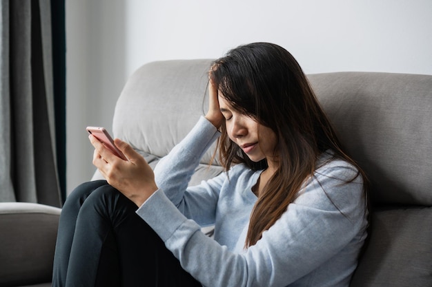 Mujer asiática joven estresada que sufre de depresión mientras sostiene el teléfono móvil en la mano sentada en el sofá en la sala de estar en casa Triste infeliz concepto decepcionado Cerrar espacio de copia