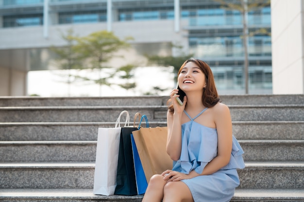 Mujer asiática joven feliz con bolsas de la compra llamando por teléfono celular y sentado en las escaleras.