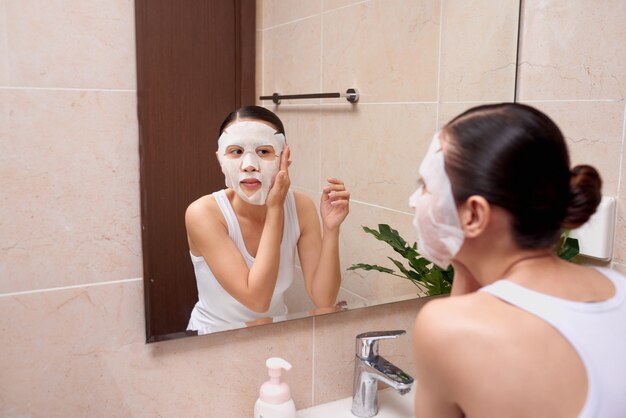 Mujer asiática joven hermosa que aplica la máscara facial cosmética en el baño.