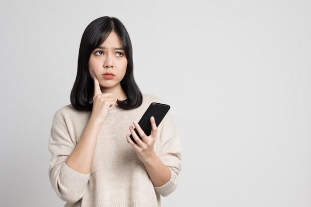 Mujer asiática joven hermosa que sostiene el smartphone que se coloca en fondo blanco aislado. Ella está mirando un espacio en blanco y está pensando en algo.