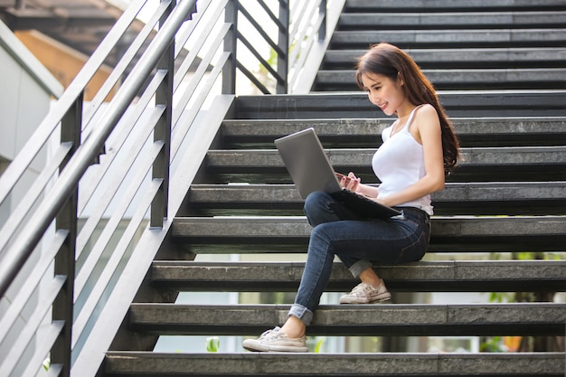 Mujer asiática joven y hermosa que trabaja con su computadora portátil mientras que se sienta en los pasos