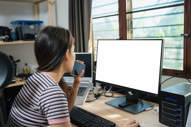 Mujer asiática joven independiente que analiza con sostener el teléfono móvil en la mesa de trabajo con el monitor vacío en casa