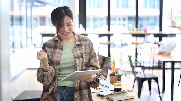 Mujer asiática joven leyendo buenas noticias agradables en tableta digital y celebrando el éxito.