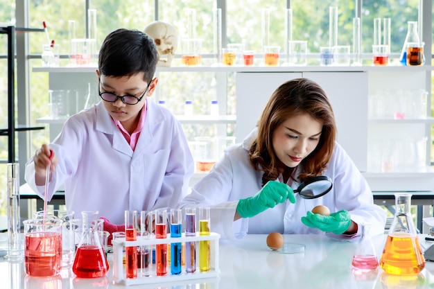 Mujer asiática joven con lupa examinando huevo de gallina mientras niño enfocado mezclando líquidos coloridos durante el experimento científico en el laboratorio de química.