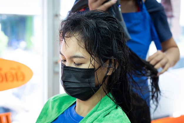 Mujer asiática joven con máscara de protección obteniendo un cabello seco con secador de pelo por peluquero en peluquería. Peluquería secando el cabello al cliente. Salón de belleza, concepto de cuidado del cabello.