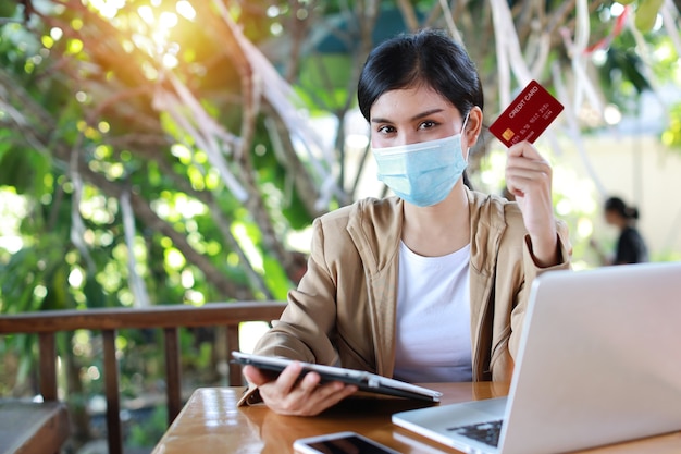 Mujer asiática joven con máscara protectora para la atención médica, sentada en la cafetería y usando el teléfono inteligente y trabajando en la computadora portátil y mostrando la tarjeta de crédito. Nuevo concepto de distanciamiento normal y social