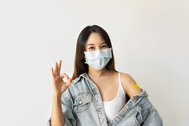 Mujer asiática joven en mascarilla protectora gesticulando bien y mostrando el brazo con vendaje de yeso después de la inyección de la vacuna Coronavirus sobre fondo blanco. Concepto de inmunización antiviral. Copie el espacio.