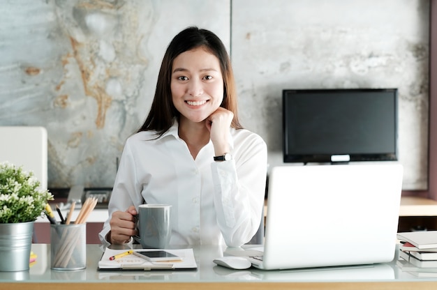 Mujer asiática joven de la oficina que trabaja con la computadora portátil y que sostiene la taza de café en el fondo de la oficina de escritorio, concepto de la forma de vida de la oficina