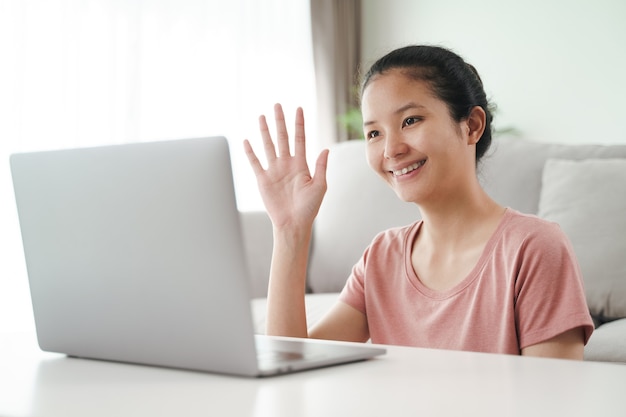 Mujer asiática joven con ordenador portátil para llamada de videoconferencia en línea agitando la mano haciendo gesto de saludo en la sala de estar.