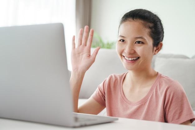 Mujer asiática joven con ordenador portátil para llamada de videoconferencia en línea agitando la mano haciendo gesto de saludo en la sala de estar.