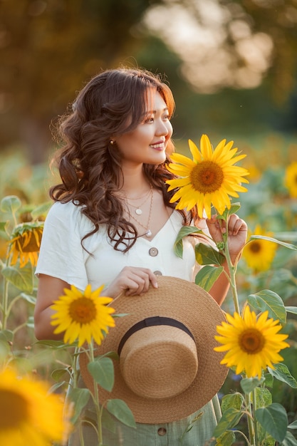 Mujer asiática joven con el pelo rizado en un campo de girasoles al atardecer. Retrato de una joven y bella mujer asiática bajo el sol. Verano.