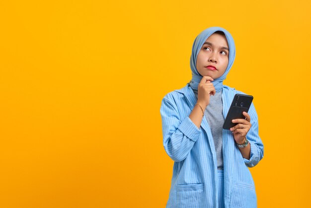 Mujer asiática joven pensativa mediante teléfono móvil con los dedos en la barbilla sobre fondo amarillo