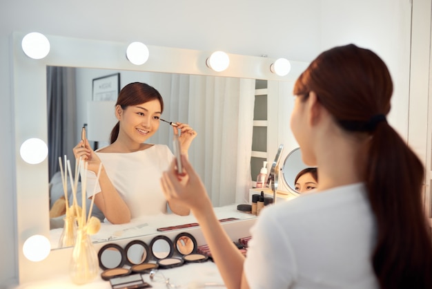 Mujer asiática joven que aplica maquillaje (pintar sus pestañas) delante de un espejo. Concéntrate en su reflejo