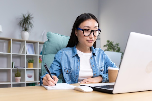 Mujer asiática joven que estudia en línea mientras está sentada en la oficina escribiendo notas de conferencias en un cuaderno usando una computadora portátil para estudiar