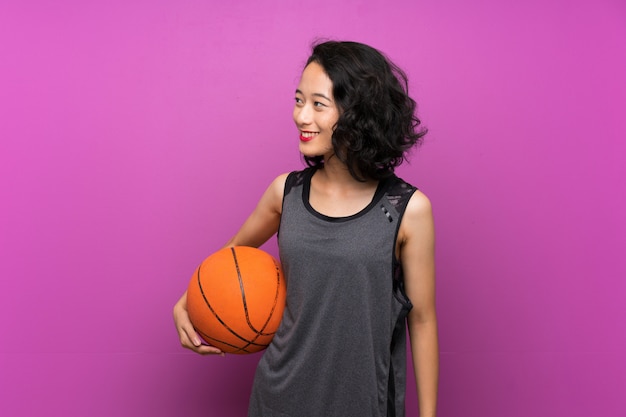 Mujer asiática joven que juega a baloncesto sobre la pared púrpura aislada