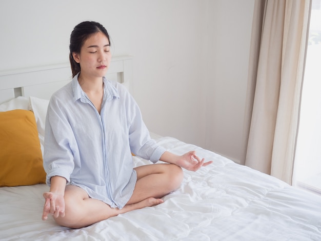 Mujer asiática joven que sienta y que practica haciendo yoga en cama