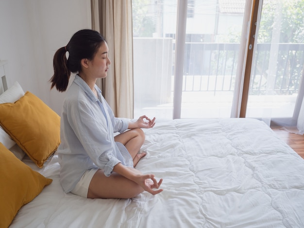 Mujer asiática joven que sienta y que practica haciendo yoga en cama