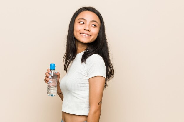 La mujer asiática joven que sostiene una botella de agua mira a un lado sonriente, alegre y agradable.