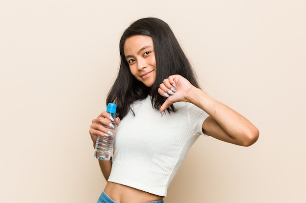 Mujer asiática joven que sostiene una botella de agua que muestra un gesto de la aversión, pulgares abajo. Concepto de desacuerdo