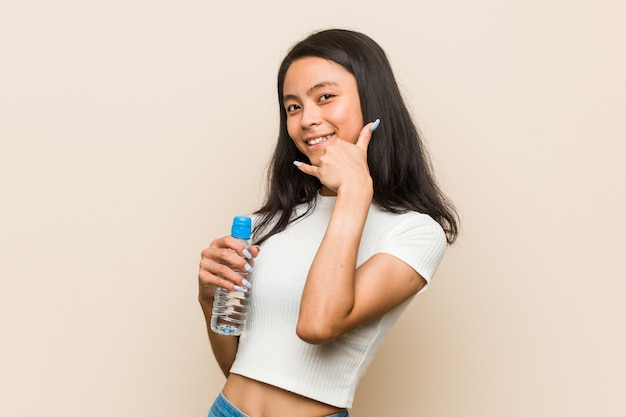 Mujer asiática joven que sostiene una botella de agua que muestra un gesto de llamada de teléfono móvil con los dedos.