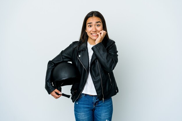 Mujer asiática joven que sostiene un casco de moto sobre la pared aislada mordiéndose las uñas, nerviosa y muy ansiosa.