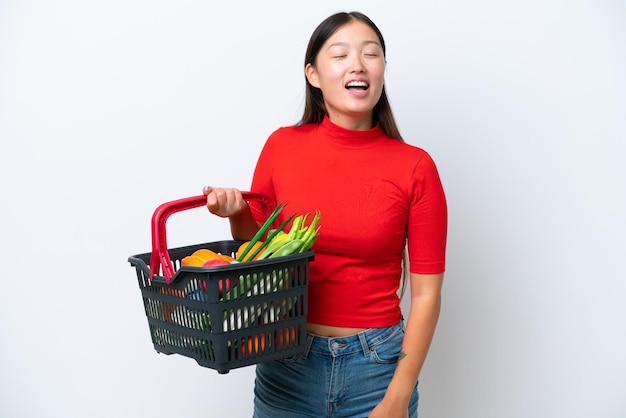 Mujer asiática joven que sostiene una cesta de la compra llena de alimentos aislado sobre fondo blanco riendo