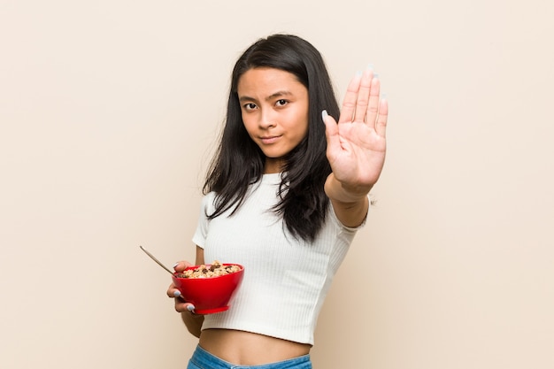 Mujer asiática joven que sostiene un cuenco de cereales de pie con la mano extendida mostrando la señal de stop, impidiéndote.