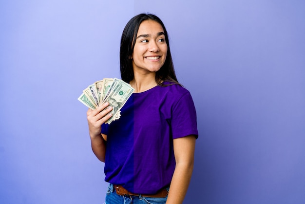 La mujer asiática joven que sostiene el dinero aislado en la pared púrpura mira a un lado sonriente, alegre y agradable.