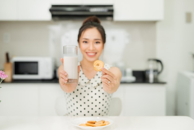 Mujer asiática joven que sostiene la galleta de la mordedura del vaso de leche en su cocina