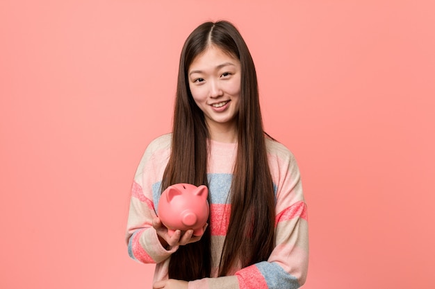 Mujer asiática joven que sostiene una hucha que sonríe confiada con los brazos cruzados.
