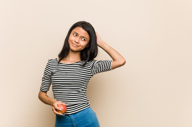 Mujer asiática joven que sostiene una manzana que toca la parte posterior de la cabeza, pensando y tomando una decisión.