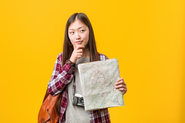 Mujer asiática joven que sostiene un mapa que mira de lado con expresión dudosa y escéptica.