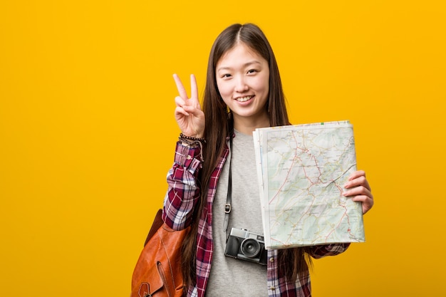 Mujer asiática joven que sostiene un mapa que muestra la muestra de la victoria y que sonríe ampliamente.