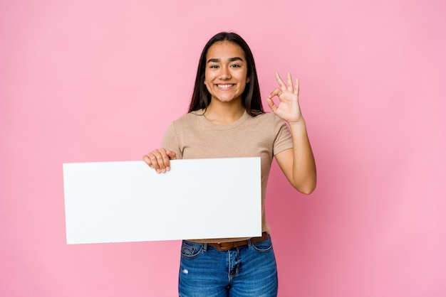 Mujer asiática joven que sostiene un papel en blanco para algo blanco sobre la pared aislada alegre y confiada que muestra el gesto aceptable.