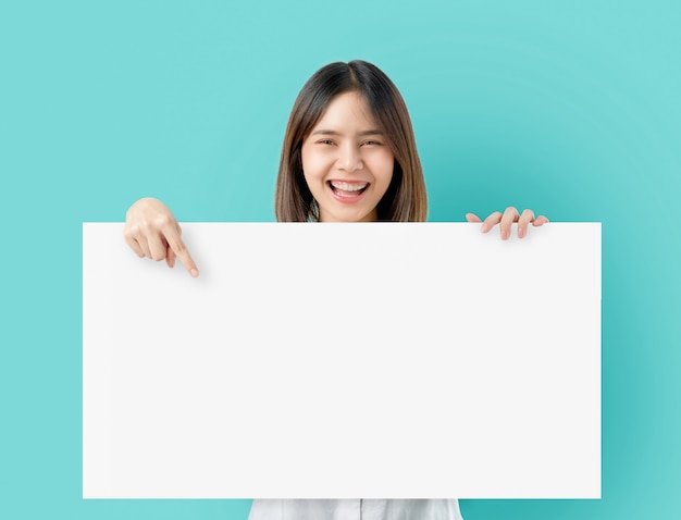 Mujer asiática joven que sostiene el papel en blanco con la cara sonriente y que mira en el azul.