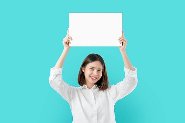 Mujer asiática joven que sostiene el papel en blanco con la cara sonriente y que mira en el fondo azul