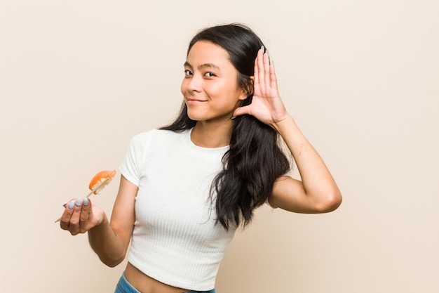 Mujer asiática joven que sostiene un pedazo de sushi tratando de escuchar un chisme.