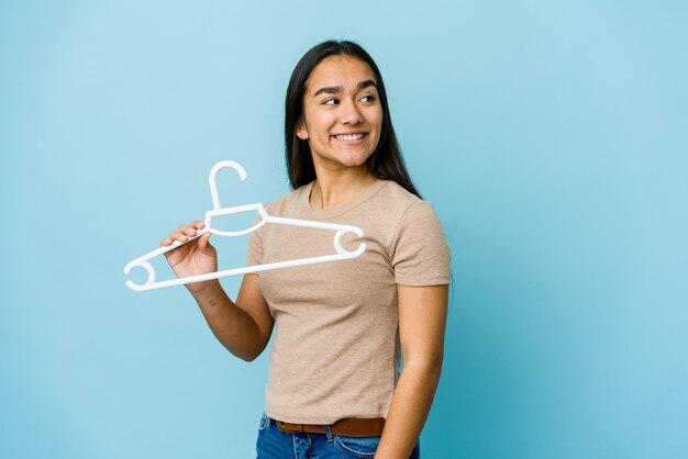 Mujer asiática joven que sostiene una percha aislada en la pared azul mira a un lado sonriente, alegre y agradable