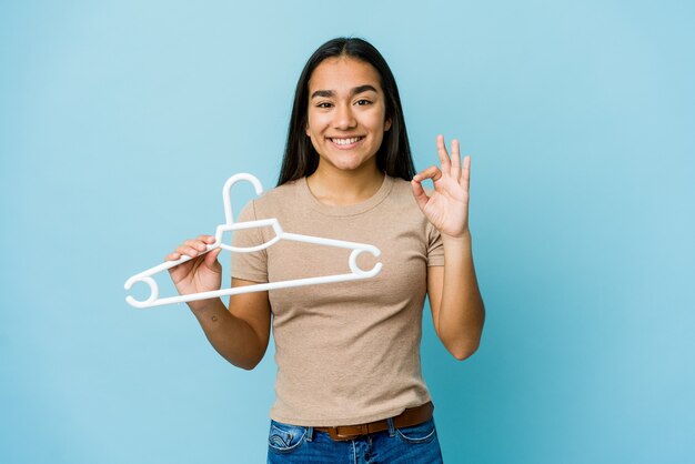 Mujer asiática joven que sostiene una percha alegre y confiada que muestra el gesto aceptable.