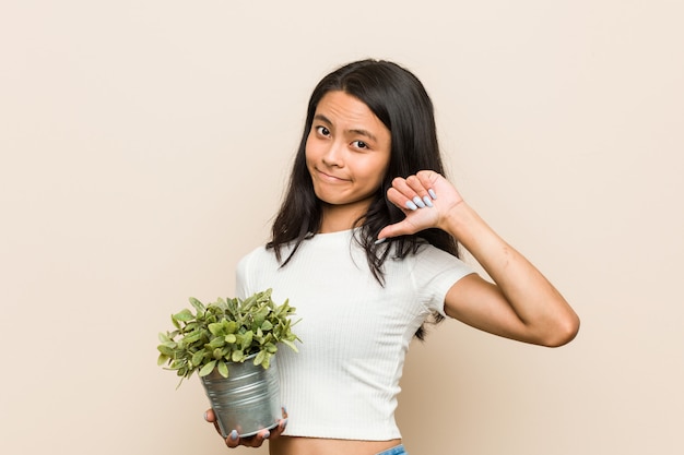 Mujer asiática joven que sostiene una planta que muestra un gesto de la aversión, pulgares abajo.